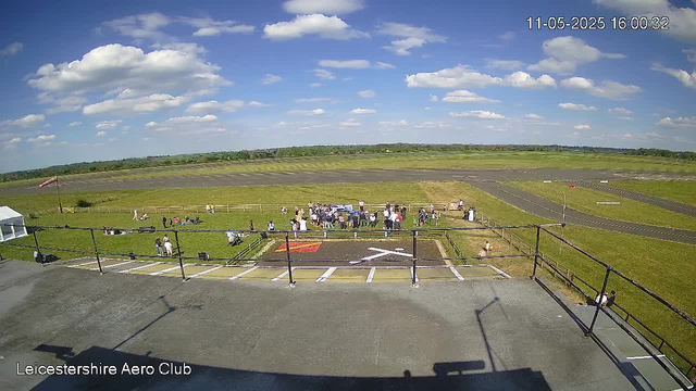 A view from a webcam shows a grassy area with a group of people gathered together, some standing and some sitting, near a low fence. To the left, there are some objects and a flagpole with a flag. In the background, there are open fields and a runway extending into the distance under a partly cloudy sky with white clouds scattered across a blue sky. The time stamp in the top right corner indicates the date and time as November 5, 2025, at 4:00 PM. The text "Leicestershire Aero Club" is visible in the bottom left corner.