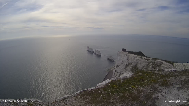 A wide view of white chalk cliffs extending into the sea, with a lighthouse and a small building on the cliff top. Several large rock formations are visible in the water near the cliffs, and the sky is partly cloudy with patches of blue. The ocean appears calm with gentle waves, and the photo was taken in the late afternoon. There is a timestamp in the bottom left corner showing "11/05/2025 17:00:25" and a website link "isleofwight.com" in the bottom right corner.