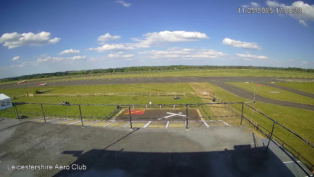 A wide view of an airfield with a clear blue sky and scattered white clouds. In the foreground, there is a rooftop or balcony with a black railing. Below, a small helipad with a white helicopter symbol is visible, along with a few parked vehicles and structures. An airplane is taking off or landing on the runway in the distance. The image has a timestamp in the top right corner, reading "11-05-2025 17:00:33," and the bottom left corner has the text "Leicestershire Aero Club."