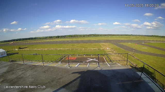 A view from a webcam showing an open grassy area with a clear blue sky and a few scattered clouds. There are patches of dark pavement and a small helipad marked with a white helicopter symbol. A fence surrounds the helipad, and a taxiway or runway extends into the distance. In the top right corner, there is a timestamp reading "11-05-2025 18:00:29." The bottom left corner has the text "Leicestershire Aero Club."