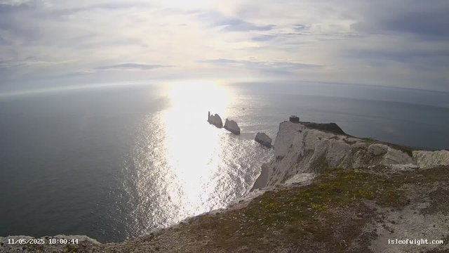 A view of white chalk cliffs on the coast, with a lighthouse visible on the edge. The sun is low in the sky, reflecting brightly on the calm sea below. There are several large rocks emerging from the water near the cliffs. The scene is partly cloudy, with the sunlight creating a shimmering effect on the water. The timestamp in the bottom left corner reads "11/05/2025 18:00:44," and the website "isleofwight.com" is visible in the bottom right corner.