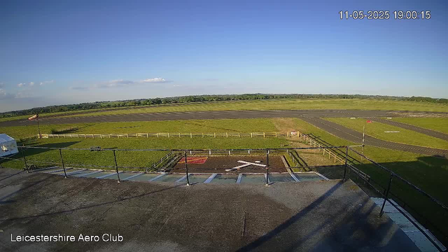 A wide view of a small airport tarmac and runway under a clear blue sky during daytime, with green fields stretching into the distance. In the foreground, there is a rooftop area with railings, some parking lines, and a small helipad marked in red. The timestamp in the top right corner reads "11-05-2025 19:00:15," and the bottom left corner indicates "Leicestershire Aero Club."