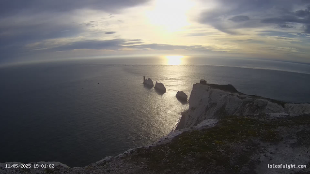 A view of the ocean at sunset with white cliffs in the foreground. Several large rock formations extend into the water, and the sky is partly cloudy with the sun low on the horizon, casting a golden light over the scene.