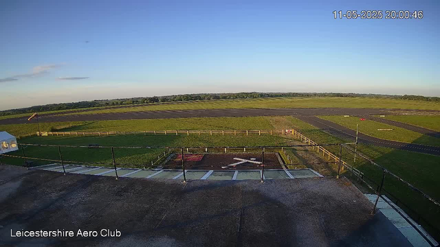 A wide view of an open grass field and a paved area at Leicestershire Aero Club during daylight, with clear blue skies. The scene includes a cleared runway area in the distance, some fencing along the perimeter, and a small building on the left side. The timestamp indicates it is evening, around 8:00 PM.