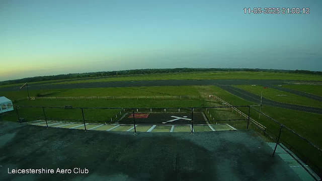 An empty rooftop helipad surrounded by a black safety railing, with a white arrow painted on the dark surface. Beyond the helipad, there is a grassy open space and an empty track or runway area. The sky is clear with a gradient from light near the horizon to darker at the top. The timestamp in the upper right corner reads "11-05-2025 21:00:12," and text in the lower left corner reads "Leicestershire Aero Club."
