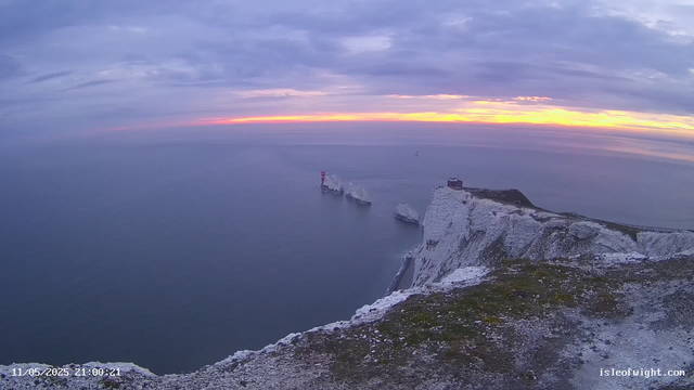 A view of white chalk cliffs overlooking the ocean at sunset, with a lighthouse on a small rock formation and a distant sailboat on the water. The sky has a mix of clouds and the colors of the setting sun, casting a warm glow on the horizon.
