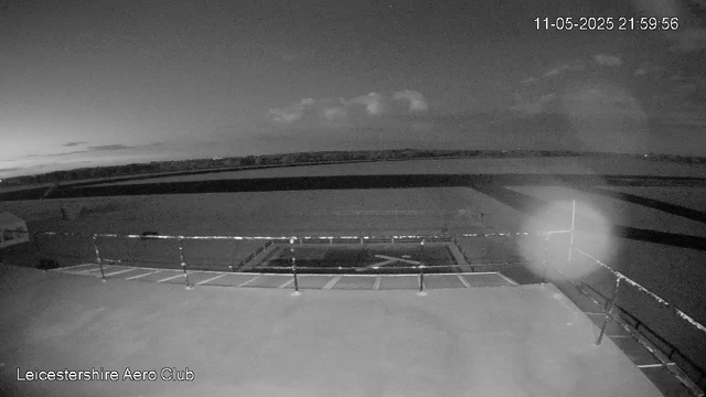 A black-and-white image from a webcam shows an empty parking lot with several parking spaces, some of which are marked with white lines. There is a gate or barrier at the front of the parking area. The background features an open sky with scattered clouds, and the horizon is visible in the distance. The timestamp in the upper right corner reads "11-05-2025 21:59:56," and the bottom left corner has the text "Leicestershire Aero Club."
