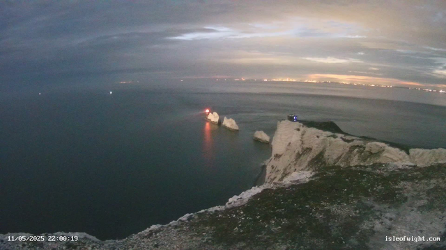 A view of white chalk cliffs overlooking the ocean at dusk or dawn. The sky is partly cloudy with a mix of dark and light clouds. Small rocks are visible on the cliff edge in the foreground. In the water, there are three prominent rocks with red and blue lights shining from vessels nearby, creating reflections on the calm ocean surface. Distant city lights can be seen on the horizon. The timestamp on the image indicates it was taken on November 5, 2025, at 10:00 PM, and the website isleofwight.com is displayed in the bottom right corner.