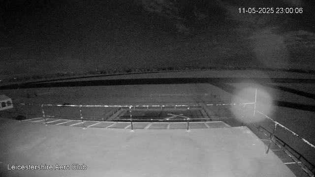 A black-and-white image of an airport tarmac at night, with empty parking spots and a runway in the distance. There is a chainlink fence in the foreground and a bright light shining from the right side. The timestamp in the top right corner reads "11-05-2025 23:00:06," and "Leicestershire Aero Club" is written at the bottom left.