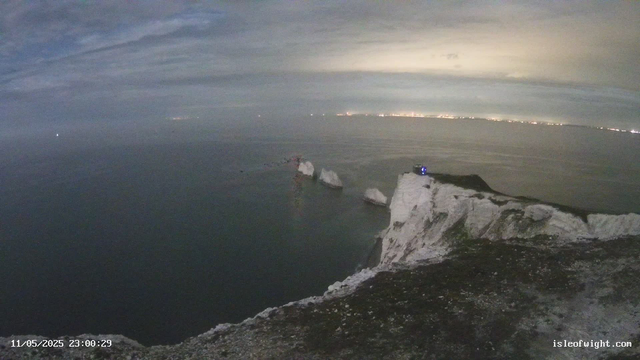 A nighttime view of white chalk cliffs overlooking the sea. The cliffs have jagged edges, and the ocean below is calm. In the distance across the water, faint lights from a distant shoreline are visible. The sky is mostly cloudy with some lighter areas. A small structure with a blue light is situated on the edge of the cliff. The timestamp at the bottom left reads "11/05/2025 23:00:29," and the bottom right has the text "isleofwight.com".