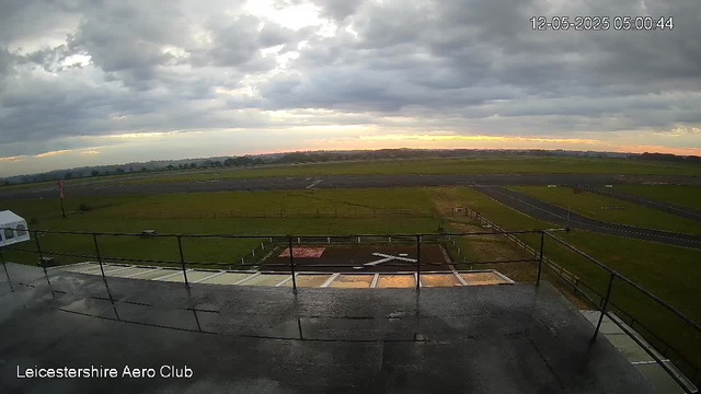 A view from a webcam during early morning at Leicestershire Aero Club. The sky is mostly cloudy with dark clouds, and there is a hint of sunlight on the horizon. The scene shows the airfield with a grassy area, runway, and taxiways in the distance. The image appears to be taken from a balcony or viewing platform with a black railing in the foreground. The timestamp indicates it is 5:00 AM on December 5th, 2025.