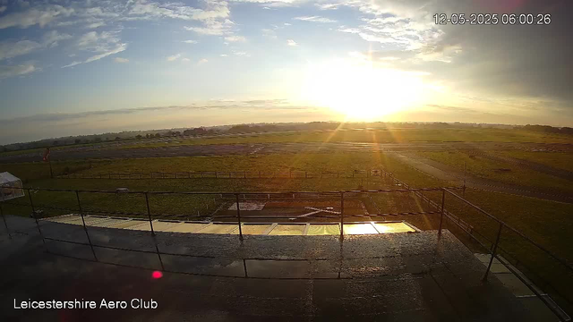 A wide view of a grassy airfield with a clear sky during sunrise or sunset, as seen from a webcam. There is a runway and some fencing in the foreground, and the sun is low on the horizon casting a warm glow across the scene. The timestamp in the upper right corner reads "12-05-2025 06:00:26," and the bottom left corner displays the text "Leicestershire Aero Club."