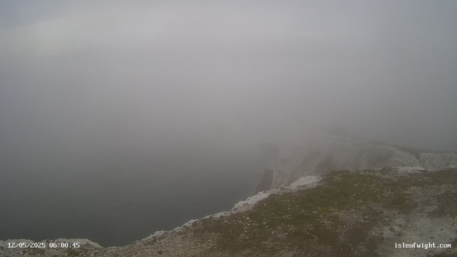A foggy, overcast scene at the edge of a rocky cliff overlooking the sea. The sky and horizon are obscured by thick fog, creating a mysterious and moody atmosphere. The rocky terrain in the foreground is covered with patches of moss and small stones. The image has a timestamp in the bottom left corner showing "12/05/2025 06:00:45" and a website address in the bottom right corner "isleofwight.com".