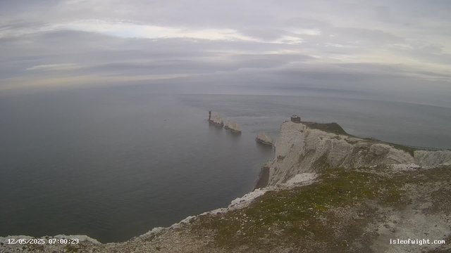 A view of white chalk cliffs rising sharply from the ocean below under a cloudy sky. The cliffs extend into the distance, with a small building and a fenced pathway near the edge. A lighthouse is visible on a separate rocky outcrop in the water, with waves gently crashing against the rocks. The scene appears to be early in the morning.