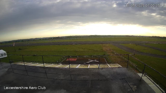 A wide view of an outdoor area at the Leicesterhire Aero Club during dawn or early morning, with a cloudy sky and a horizon with fields and distant trees. The foreground shows a helipad with a white "H" marking and a fenced perimeter, along with some open grassy fields and paved pathways extending into the distance. A timestamp in the upper right corner indicates the date and time as 12-05-2025 07:00:25.