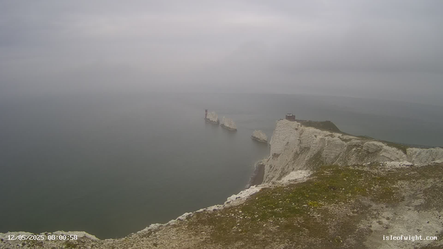 A view of white chalk cliffs extending into the ocean on a foggy day. Several large chalk rock formations are visible offshore. There is a lighthouse on the cliff's edge towards the left side of the image, and a building near the lighthouse. The ground in the foreground is uneven and covered with patches of grass and small stones. The sky is overcast, creating a moody atmosphere.