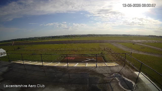 An outdoor scene showing a wide view of a grassy airfield with a runway in the distance under a partly cloudy sky. There is a railing and a small platform in the foreground, with a sign and some structures nearby. The timestamp in the top right corner reads "12-05-2025 08:00:57," and the bottom left corner has the text "Leicestershire Aero Club."