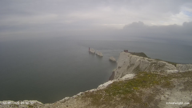A view of white chalk cliffs extending into the sea, with three large rock formations just offshore. The sky is overcast with gray clouds, and the water appears calm. There is a small building or structure near the edge of the cliffs.