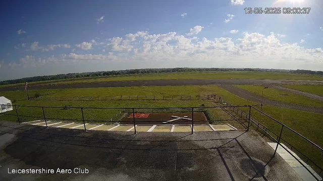 A view from a webcam showing an open grassy airfield with a clear blue sky overhead and scattered clouds. In the foreground, there is a metal railing and a small parking area with white marked spaces. To the left, part of a white tent or canopy is visible, and towards the distance, the grassy runway extends into the horizon. Rolling fields and distant trees are visible in the background. The timestamp indicates the date as December 5, 2025, at 9:00 AM. The text "Leicestershire Aero Club" is displayed in the bottom left corner.