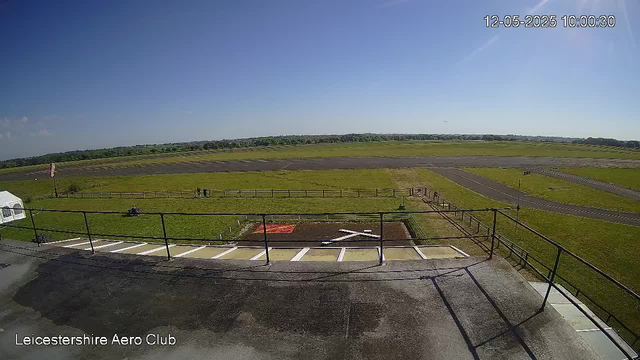This image shows a view from a webcam at Leicestershire Aero Club on a clear day. The foreground includes a platform with railing, a small parking area with white lines and a tent on the left. Beyond the platform, there is a grassy field with a few people standing or sitting near a fence. The background features a wide open space with a paved pathway and several runways or taxiways, and distant tree lines under a bright blue sky with a few small clouds. The timestamp in the upper right corner reads "12-05-2025 10:00:30."