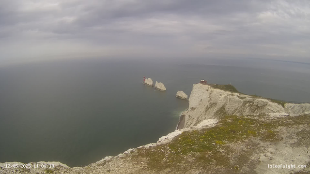 A view of steep white chalk cliffs overlooking a calm, gray sea under a cloudy sky. Several white chalk stacks rise from the water near the cliffs, with a small red lighthouse on one of the stacks. Atop the main cliff, there is a small building or lookout point, and a pathway runs along the edge of the cliff. The foreground shows the uneven, mossy edge of the cliff.