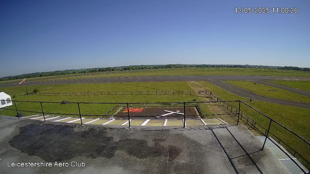 A view from a webcam at Leicesterhire Aero Club shows a clear, sunny day with a bright blue sky. In the foreground, there is a section of paved area with a metal railing, parking spaces, and a small marked pad with an airplane symbol. The middle ground features a grassy field with a wooden fence, and beyond that, a large open grassy expanse with a few paved taxiways or runways. The horizon reveals a distant line of trees or low hills. The image is timestamped "12-05-2025 11:00:26" in the top right corner, and the bottom left corner has the text "Leicestershire Aero Club."