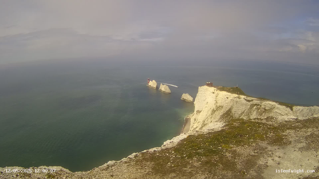 A panoramic view of white chalk cliffs overlooking the ocean, with several large rocks emerging from the water near the base of the cliffs. There is a lighthouse on one of the smaller rocks at the shoreline, and a building or structure at the top of the main cliff. The sky is mostly cloudy with patches of blue, and the overall scene is calm and expansive.