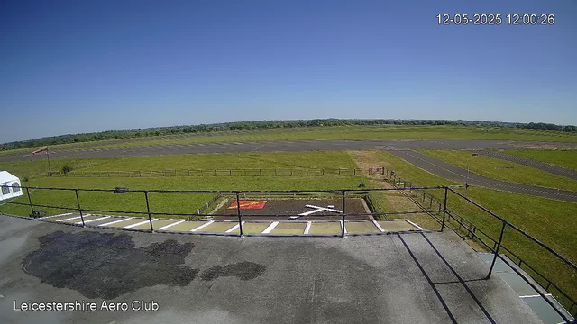 A wide view of an open airstrip and grassy fields under a clear blue sky. In the foreground, there is a parking area with white lines and a black railing. A small section of the tarmac with a marked helicopter landing spot is visible, along with a pathway leading from the parking area towards the open field. The landscape stretches out into the distance with no buildings or trees nearby. The image is timestamped "12-05-2025 12:00:26" and labeled "Leicestershire Aero Club" in the bottom left corner.