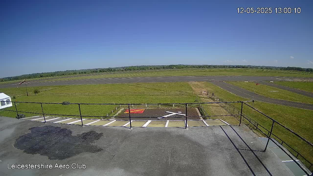 A view from a webcam showing an open grassy area and an empty tarmac runway under a clear blue sky. In the foreground, there is a parking area with white lines and a metal railing, along with some dark patches on the asphalt. The background extends to a flat horizon with distant trees. The top right corner displays a timestamp "12-05-2025 13:00:10" and the bottom left corner has the text "Leicestershire Aero Club."