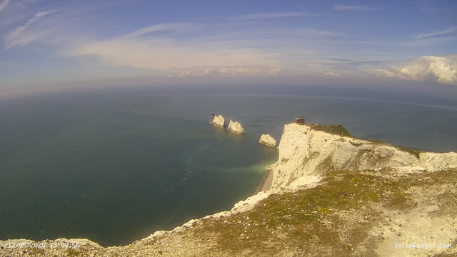 Aerial view of white chalk cliffs extending into the sea, with a small structure on top of the nearest cliff. Three large rock stacks are visible in the water beyond the cliffs, with a lighthouse on one of them. The sky is mostly clear with some clouds and the ocean appears calm. The date and time stamp in the bottom left corner reads "12/05/2025 13:00:56".