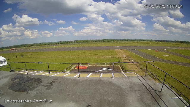 An outdoor view from a webcam at Leicesterhire Aero Club shows a small parking area with marked spaces, a grassy airfield with taxiways, and a partly cloudy sky with white clouds scattered across a blue background. There is a black fence surrounding the parking area and a white tent visible on the left side. The timestamp in the upper right corner reads "12-05-2025 14:00:04".