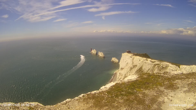 Aerial view of white chalk cliffs along the coastline under a partly cloudy sky. The sea is calm with a boat creating a wake as it moves near the cliffs. There is some greenery and moss on the edge of the cliffs. A timestamp is visible in the lower left corner.