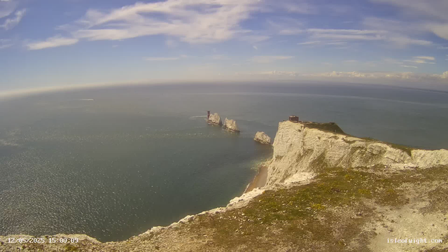 A panoramic view of white chalk cliffs rising sharply from the sea, with a grassy top and some buildings on the cliffs. In the distance, the sea stretches to the horizon under a partly cloudy sky, with a few small ships or boats visible on the water. The date and time stamp in the bottom left corner indicates "12/05/2025 15:00:09," and the scene is illuminated by bright sunlight.