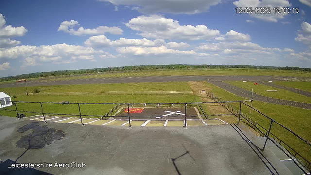 A clear view of a sunny day at Leicestershire Aero Club shows a paved area with a safety railing in the foreground, leading to an open grassy airfield with a small runway. The sky is bright blue with scattered fluffy white clouds, and distant green hills stretch across the horizon. A timestamp in the top right corner reads "12-05-2025 15:00:15".