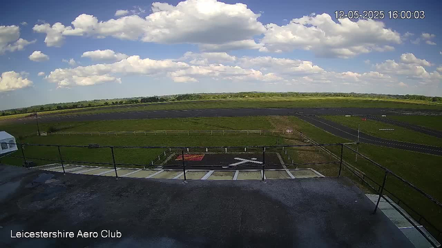 An outdoor scene showing a clear sky with scattered white clouds over open green fields and a racetrack. In the foreground, there is a rooftop or a viewing platform with a safety railing and marked helipad. The image is from a webcam at Leicestershire Aero Club, with the date and time displayed in the top right corner as "12-05-2025 16:00:03."