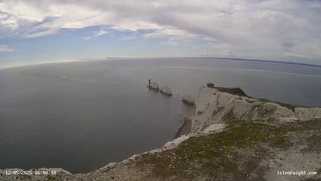 A panoramic view of white chalk cliffs and rock formations overlooking the sea. The sky is partly cloudy with patches of blue, and the water appears calm. There are a few small buildings on top of the cliffs and a winding path along the edge. The image is timestamped December 5, 2025, at 16:00:48.
