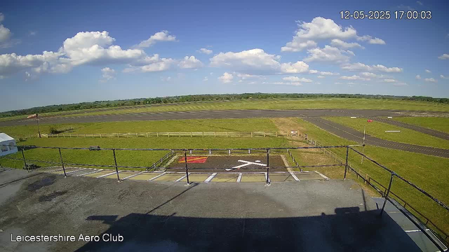 A view from a webcam showing an open, grassy airfield under a bright blue sky with scattered white clouds. In the foreground, there is a small parking area with parking spaces marked, and a closed gate or barrier. The background reveals a runway with a red vehicle parked on it, fencing around the airfield, and distant trees on the horizon. The image is timestamped December 5th, 2025, at 5:00 PM, and the bottom left corner displays the text "Leicestershire Aero Club."