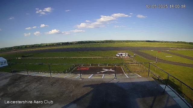 View of an open grassy airfield with a small white van parked near a fenced area. A person is standing next to the van. The sky is clear with some scattered clouds, and the horizon shows distant trees and fields. The image is timestamped "12-05-2025 18:00:18" and has the label "Leicestershire Aero Club" at the bottom left.