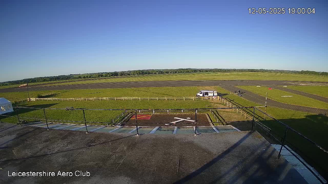 A wide view of a grassy airfield with a paved runway extending into the distance under a clear blue sky. In the foreground, there is a flat rooftop with black railings, on which a few shadows fall. To the left, a white tent is visible. Near the center, there is a white van and a motorcycle parked on a small paved area, with a person standing nearby. A flag on a pole is seen to the left of the van. The background shows open fields and trees along the horizon. The image has a timestamp in the upper right corner indicating the date and time as December 5, 2025, at 19:00:04. Text in the bottom left corner reads "Leicestershire Aero Club."