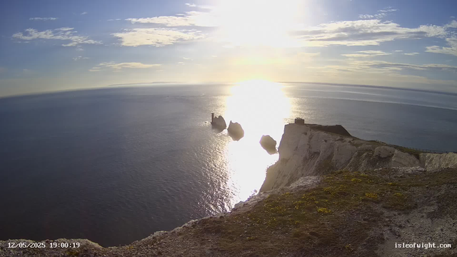 A panoramic view of a seaside cliff at sunset, with the sun low on the horizon casting bright reflections on the water below. The cliff has a white, chalky appearance with patches of green moss or lichen, and a small structure or building is visible near the edge. Three rocky sea stacks rise from the water close to the cliff, with the largest one featuring a small tower or lighthouse on top. The sky is partly cloudy with scattered clouds allowing sunlight to stream through.