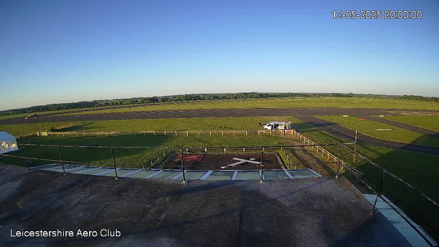 A wide-angle view from a webcam showing a clear sky and an open grassy area with some paved pathways. In the foreground, there is a rooftop with black railings and white rectangular markings. A white vehicle is parked near a fenced area with green grass, and there is a small tent on the left side. The landscape extends into fields and distant trees on the horizon. The image is timestamped "12-05-2025 20:00:00" and has the text "Leicestershire Aero Club" at the bottom left corner.