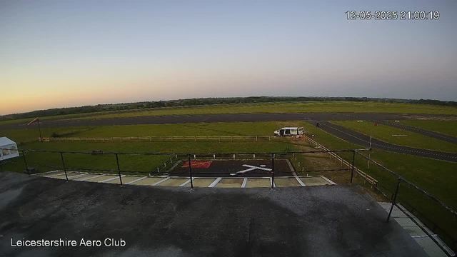 A view of an airfield at sunset or sunrise with a clear sky and open green fields in the background. In the foreground, there is a small paved area with white lines, a black fence, and a helicopter landing pad marked with a white 'H' and a red square. A white vehicle is parked near the edge of the grassy area. The timestamp in the top right corner reads "12-05-2025 21:00:19." The bottom left corner has text that says "Leicestershire Aero Club."