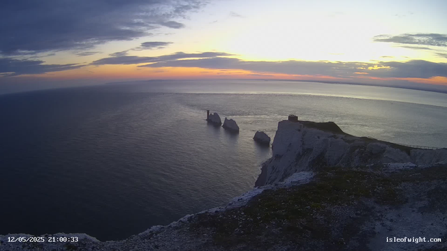 A wide view of white chalk cliffs overlooking a calm ocean at sunset. The sky has clouds with hues of blue, orange, and yellow. There are three large sea stacks extending from the cliffs into the water, with one having a small lighthouse on top. A pathway runs along the cliff edge on the right side of the image. The date and time are shown in the bottom left corner, and a website URL is in the bottom right corner.