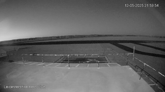 A black-and-white image of an outdoor area at night, showing a flat paved surface enclosed by a metal railing. In the background, a vast open landscape stretches towards the horizon under a dark sky. The date and time stamp in the upper right corner reads "12-05-2025 21:59:54," and the text in the lower left corner reads "Leicestershire Aero Club."