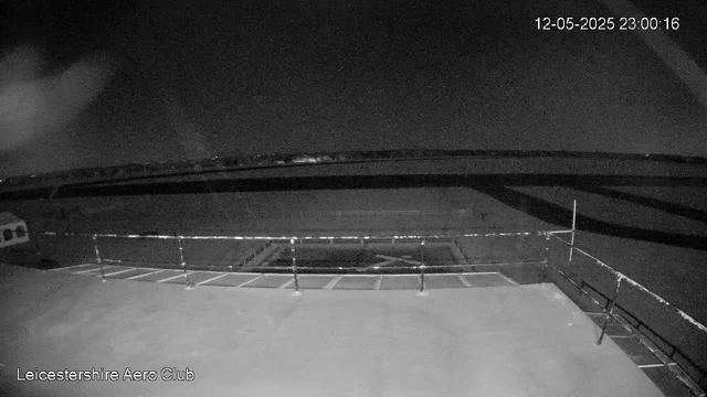 A black and white webcam image shows a view of an empty outdoor area at night, with a fenced section in the foreground and a dark sky above. The date and time displayed on the top right corner read "12-05-2025 23:00:16." The bottom left corner has the label "Leicestershire Aero Club."