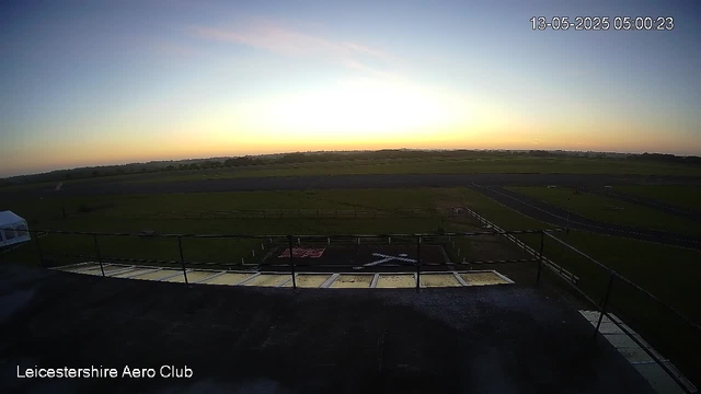 A view of an open field and runway at sunrise or sunset, with a clear sky and a bright horizon on the horizon. The scene is captured from a webcam at the Leicestershire Aero Club, with the timestamp 13-05-2025 05:00:23 displayed in the upper right corner.
