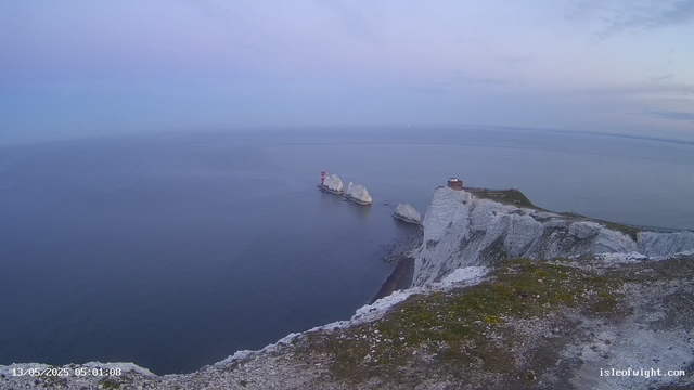 A view of white chalk cliffs over the ocean at dawn or dusk. The cliffs extend into the sea, with some large rocks or small islands nearby, topped with a small building or lighthouse. The sky is mostly cloudy with soft pastel colors. The image has a timestamp of 13/05/2025 05:01:08 and a website watermark "isleofwight.com".