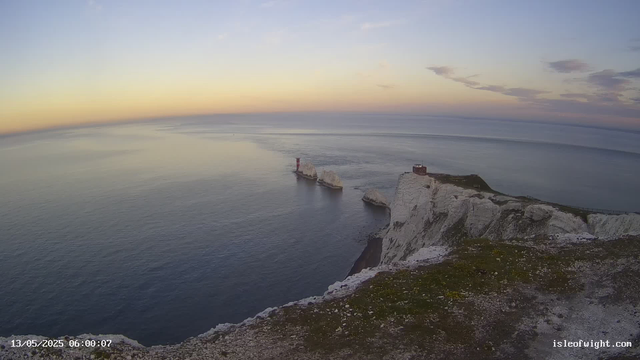 A view from a webcam showing white chalk cliffs on a coastline during sunset, with a lighthouse perched on the edge of the cliffs. The sky has soft pastel colors, and the calm sea reflects the light, extending to the horizon. There is a timestamp in the bottom left corner that reads "13/05/2025 06:00:07," and a website URL "isleofwight.com" in the bottom right corner. The overall scene is tranquil and scenic.