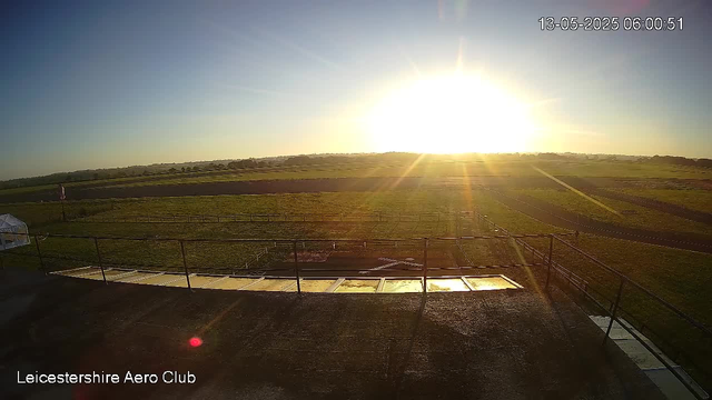 The image shows a sunrise or sunset over an open field with green grass, sectioned by fences and pathways. The sky is clear, with the sun low on the horizon, casting bright light and long shadows across the landscape. In the foreground, part of a black railing and a small section of a transparent structure can be seen. The words "Leicestershire Aero Club" are written in the bottom left corner, and the timestamp in the top right corner indicates the date and time as 13-05-2025 at 06:00:51.