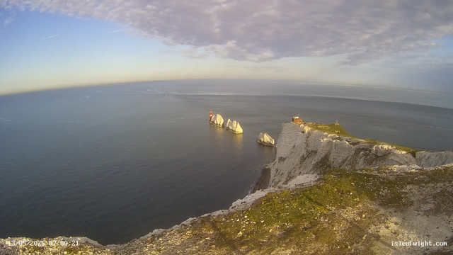 Aerial view of white chalk cliffs overlooking the ocean with large white rock formations in the water. There is a small building and a lighthouse on the grassy top of the cliff, with the sky partly cloudy and the horizon stretching far into the distance. The scene appears to be during the early morning or late evening based on the lighting. The image includes a timestamp in the bottom left corner and the website "isleofwight.com" in the bottom right corner.